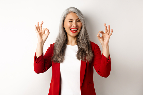 Asian professional businesswoman showing OK signs and winking, smiling pleased, assure or recommend something, standing against white background.