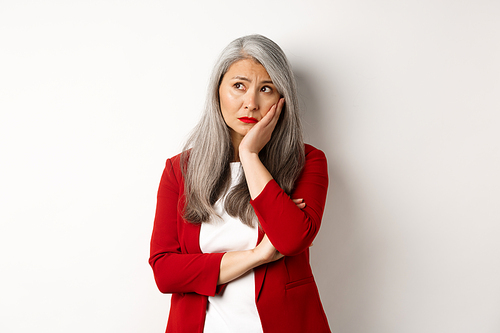 Troubled and sad asian female entrepreneur looking at upper left corner, thinking with upset face, standing over white background.