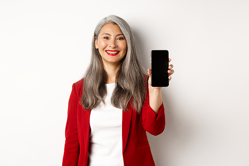 Elderly businesswoman in red blazer, showing blank smartphone screen and smiling, demonstrate mobile app, white background.