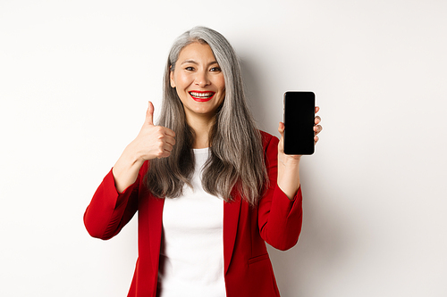 Satisfied asian elderly businesswoman showing blank smartphone screen and thumb-up, praising online promotion or company app, standing over white background.