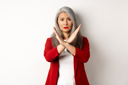 Asian senior woman in red blazer making cross gesture, pleading you stop, disagree and disapprove something, standing over white background.
