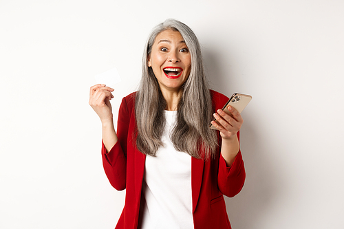 Excited asian businesswoman in red blazer, holding plastic credit card and smartphone, smiling happy at camera, standing over white background.