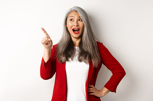 Surprised senior asian lady pointing upper left corner, checking out promo offer and smiling amazed, standing in red blazer over white background.