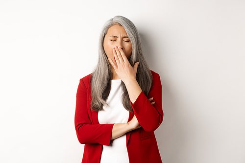 Tired or bored asian businesswoman yawning, covering mouth with hand, standing exhausted over white background. Copy space