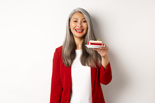 Celebration and holidays concept. Smiling asian businesswoman congratulate employees, giving plate with sweet cake and looking happy, standing over white background.