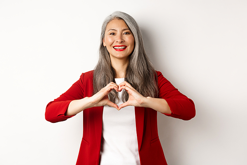 Beautiful asian mature woman in red blazer and makeup, showing heart sign and smiling, I love you gesture, standing over white background.