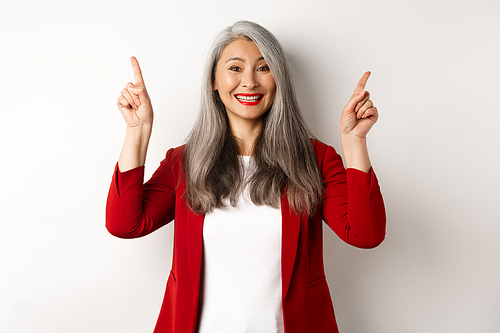 Happy mature woman in red blazer and makeup, smiling and showing advertisement on top, pointing fingers up at logo, white background.