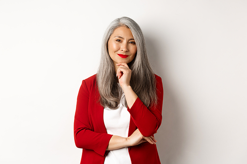 Business concept. Asian mature businesswoman smiling pleased, looking thoughtful, having an idea, standing in red blazer over white background.