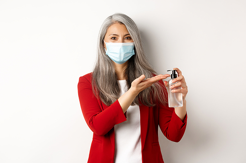 Covid, pandemic and business concept. Asian female manager in medical mask using hand sanitizer and smiling at camera, standing with antiseptic over white background.