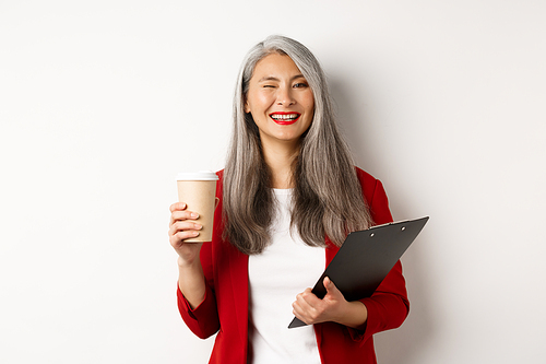 Successful asian businesswoman drinking coffee and winking cheerful at camera, standing with clipboard, wearing red blazer, white background.