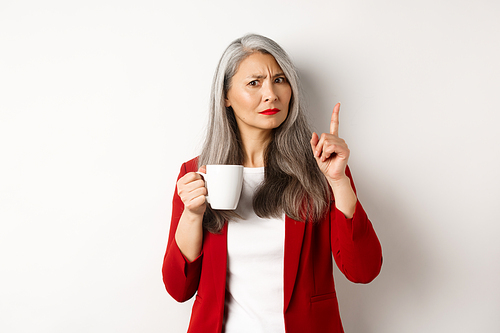 Business people concept. Displeased asian businesswoman drinking coffee and shaking finger with disapproval, prohibit something, standing over white background.