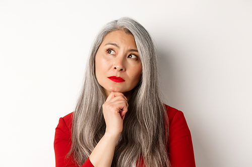 Close up of thoughtful asian business woman looking at upper left corner, thinking or making choice, standing over white background.