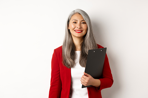 Successful asian senior business woman holding clipboard, wearing red blazer and lipstick at work, smiling at camera, white background.