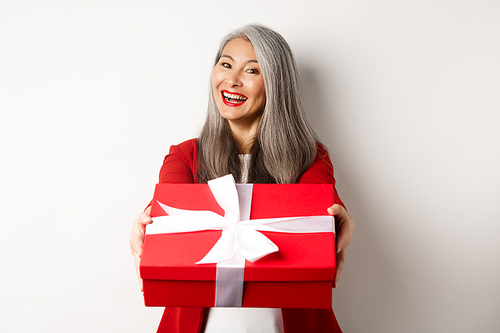 Happy holidays concept. Cheerful asian senior woman giving you red gift box and smiling, congratulating with valentine day, standing over white background.