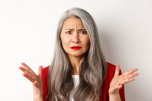 Close up of confused asian female manager with grey hair, wearing red blazer and makeup, spread hands sideways and staring puzzled at camera, white background.