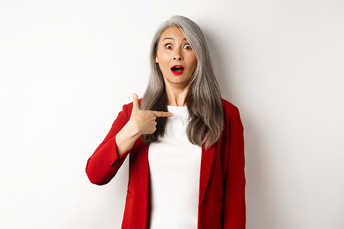 Surprised asian woman with grey hair, pointing at herself and gasping confused, standing over white background.