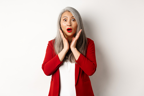 Business people. Asian senior businesswoman in red blazer and makeup gasping, looking surprised at camera, standing over white background.