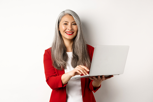 Business. Senior woman working on laptop, wearing office outfit and smiling, standing over white background.