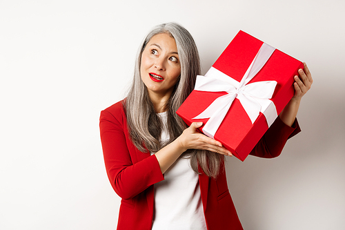 Elegant asian senior woman shaking gift box, trying to guess what inside present, standing over white background. Holidays and celebration concept.