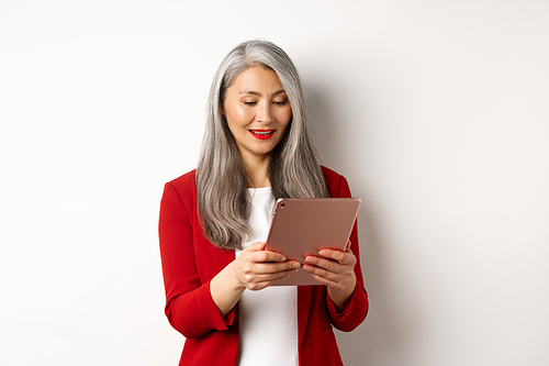 Business. Successful senior businesswoman working with digital tablet, reading screen and smiling, standing in elegant red blazer over white background.