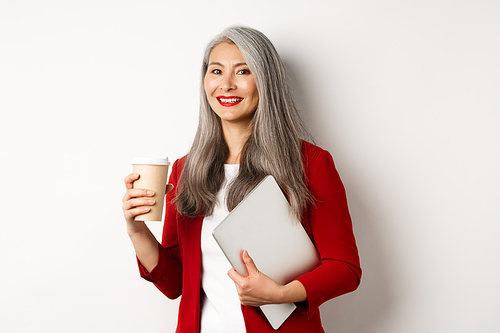 Business. Successful asian businesswoman with grey hair, wearing red blazer, drinking coffee and standing with laptop in hand, white background.