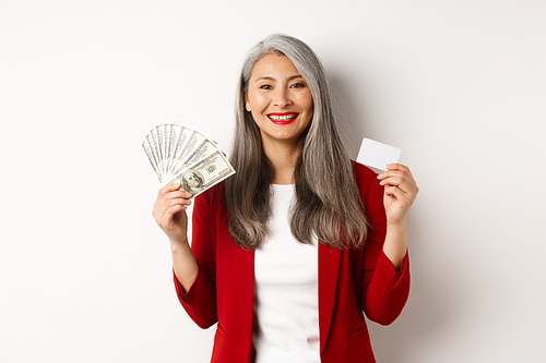 Successful asian senior businesswoman showing money in dollars and plastic card, smiling happy at camera, wearing red blazer and make-up.