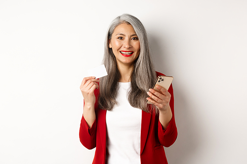 Asian middle-aged woman using plastic credit card and smartphone to shop online, standing over white background.