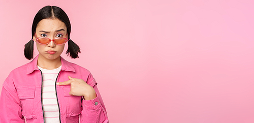 Portrait of asian girl looks confused and points at herself, perplexed face, stares with disbelief at camera, stands over pink background.