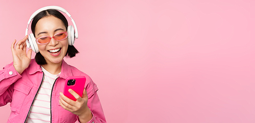 Stylish asian girl dancing with smartphone, listening music in headphones on mobile phone app, smiling and laughing, posing against pink background.