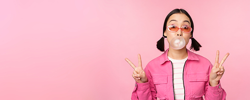 Beautiful korean girl in sunglasses, blowing bubblegum bubble and showing peace signs, standing over pink background. Copy space