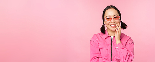 Close up portrait of stylish asian woman in sunglasses, smiling, looking flattered, laughing coquettish, standing over pink background. Copy space