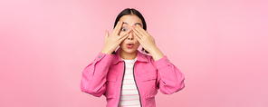 Portrait of asian girl peeks with excitement through fingers, covers eyes, seeing surprise, standing over pink background. Copy space