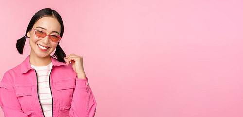 Beautiful asian girl in sunlgasses, smiling at camera, posing against pink studio background.
