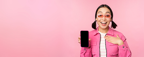 Image of surprised girl showing mobile phone app screen, smartphone display, application interface, standing against pink background.