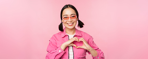 Lovely korean female model in trendy sunglasses, shows heart, care sign, I love you gesture, stands over pink background.