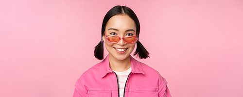 Close up of stylish korean girl in sunglasses, smiling happy, posing against pink background. People face concept.