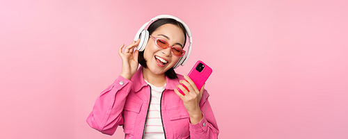 Stylish asian girl dancing with smartphone, listening music in headphones on mobile phone app, smiling and laughing, posing against pink background.