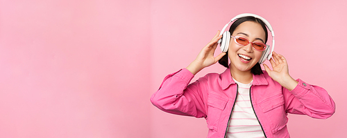 Dancing stylish asian girl listening music in headphones, posing against pink background.