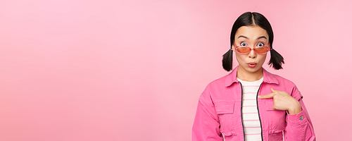 Close up portrait of asian girl looks surprised, points at herself with disbelief, being chosen, stands over pink background. Copy space