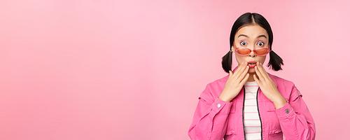 Portrait of young asian woman in stylish sunlgasses, looking surprised, disbelief reaction, standing over pink background. Copy space