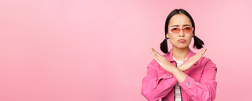 Portrait of korean girl in stylish sunglasses, sulking disappointed, showing stop, rejection gesture, cross sign, standing over pink background.