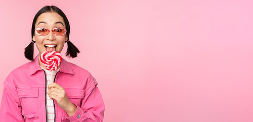 Stylish korean girl licking lolipop, eating candy and smiling, standing in sunglasses against pink background.