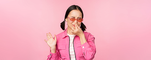 Stinky. Asian girl in sunglasses and stylish outfit, shuts her nose from disgust, dislike bad smell, standing over pink background.