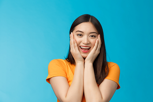 Flirty cute asian girl look surprised, express admiration and joy, touch cheeks, blushing coquettish smiling receive good news, stand blue background happily react amazed and excited.