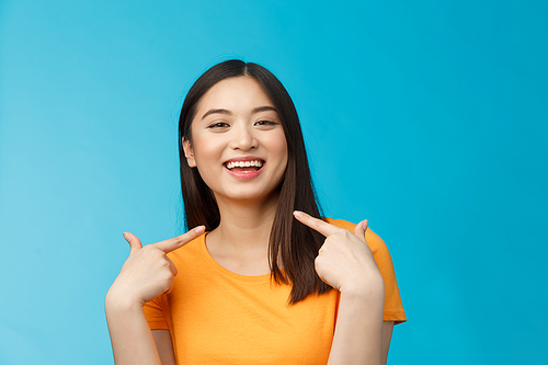 Close-up proud boastful cute asian girl telling story pointing herself, smiling broadly, bragging new haircut, standing pleased, delighted tell big news, stand blue background upbeat.