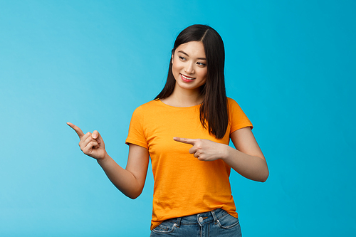 Tender lovely asian girl with dark haircut look curiously pointing left, interested in new product in store, stand blue background amused, entertained, observe exhibition, wear yellow t-shirt.