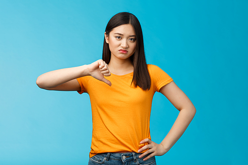 Moody unsatisfied cute asian girl picky, disliking bad restaurant food smirking displeased, grimacing disappointed show thumb down, shaking head negative judgement, stand blue background.