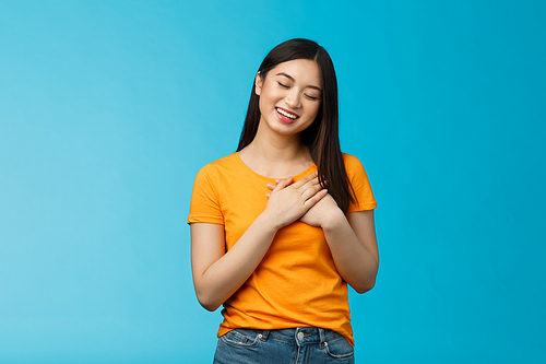 Lovely asian woman with dark haircut close eyes press hands heart gently smiling, tilt head cute cherish heartwarming memories, daydreaming, imaging beautiful dream, stand blue background.