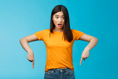 Impressed asian girl drop jaw shocked look camera surprised pointing down indicaring bottom advertisement, gasping open mouth astonished curious about interesting opportunity, blue background.