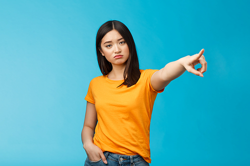 Go away from here. Serious-looking arrogant displeased bossy asian girl showing way-out, pointing sideways index finger look camera demand leave, stand blue background ignorant. Copy space